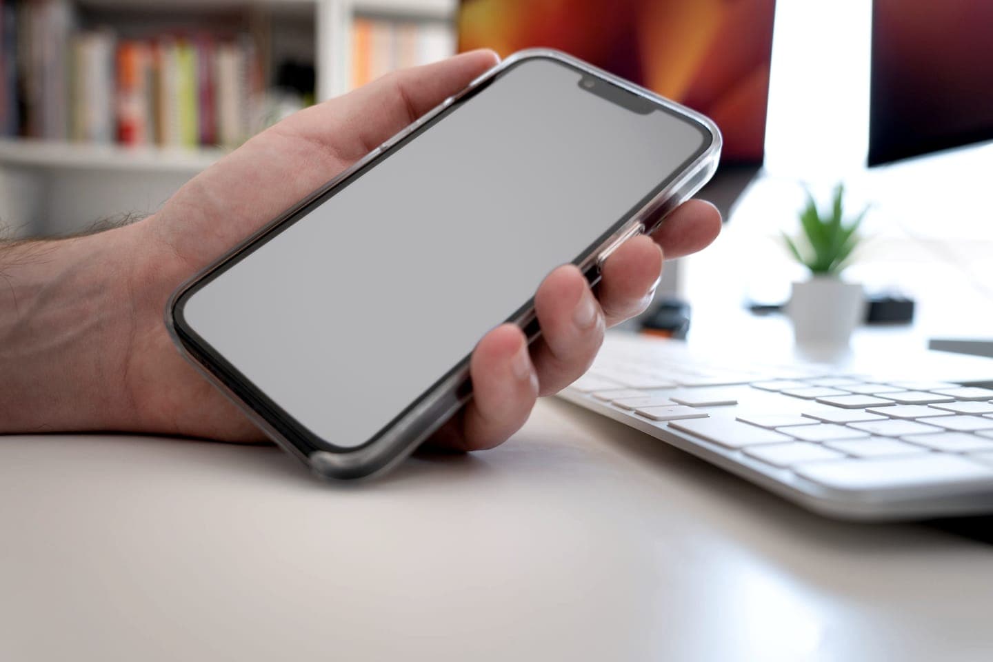 A hand holding a smartphone above a desk with a keyboard